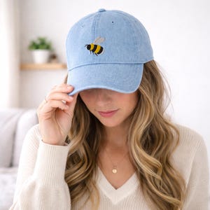Peut inclure: Casquette de baseball bleu clair avec une broderie d'abeille. L'abeille est jaune, noire et blanche. La casquette est en tissu doux de type denim et possède une visière incurvée.