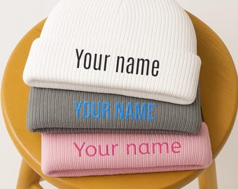 May include: Three ribbed knit beanies in white, grey, and pink are stacked on a wooden stool. Each beanie has embroidered text: "Your name" in black on the white and pink beanies, and "YOUR NAME" in blue on the grey beanie.