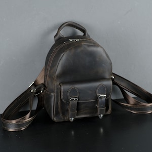 Può includere: Uno zaino piccolo in pelle marrone con due tasche anteriori e fibbie argentate. Lo zaino ha spalline regolabili.