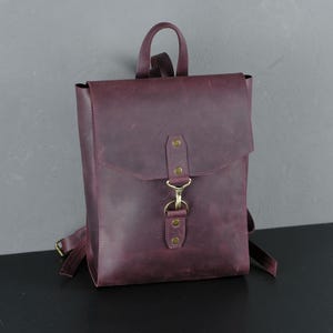 May include: A deep purple leather backpack with a gold buckle and a flap closure. The backpack has a simple design and is made of high-quality leather.