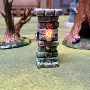 May include: A miniature stone tower with a lit torch, perfect for tabletop gaming. The tower is gray with green accents, and the torch emits a warm orange glow. Two miniature trees are in the background, set on a green surface.