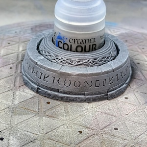 May include: A gray, 3D-printed paint pot holder with the text "CITADEL COLOUR" on the paint pot. The holder has a decorative design with etched lettering around the base. The holder is designed to hold a paint pot.