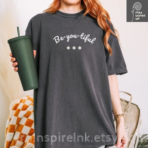 Könnte beinhalten: Ein überdimensioniertes T-Shirt in Dunkelgrau mit dem Text "Be-you-tiful" in weißen Buchstaben und drei weißen Gänseblümchen darunter.