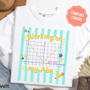 May include: A white t-shirt featuring a graphic with light blue vertical stripes and a mathematical coordinate plane showing the absolute value function y = |x|. The text reads Working on my Abs in yellow, accompanied by colorful stars and a pencil.