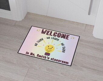 May include: Rectangular doormat with a black border and a light-colored background. The mat features the word "WELCOME" in maroon and black, along with the phrases "We Think, We Learn, We Shine." A smiling lightbulb graphic is in the center, with the text "in Ms. Smith's classroom."