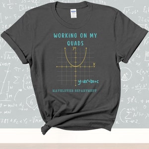 May include: Dark grey t-shirt featuring the text "WORKING ON MY QUADS" above a yellow parabola graph. The equation "y=ax²+bx+c" and the text "MATHLETICS DEPARTMENT" are below the graph.