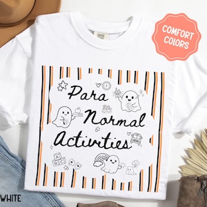 May include: A white t-shirt featuring a graphic with black and orange vertical stripes. The design includes the text "Para Normal Activities" in black script, surrounded by illustrations of ghosts, a school bus, books, and alphabet blocks.