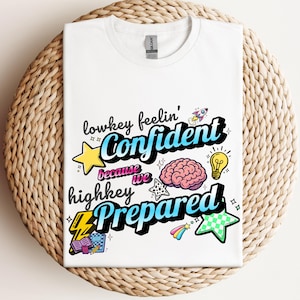 May include: White t-shirt with the text "lowkey feelin' Confident because we highkey Prepared" in a colorful, cartoonish design. The design includes a star, brain, lightbulb, and other graphics. The shirt is folded on a woven surface.