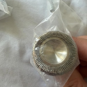 May include: A silver-colored, circular knob with a ridged outer edge and a smooth, central disc. The knob is encased in a clear plastic bag, suggesting a new or unused condition. The design is simple and functional, suitable for furniture or cabinetry.