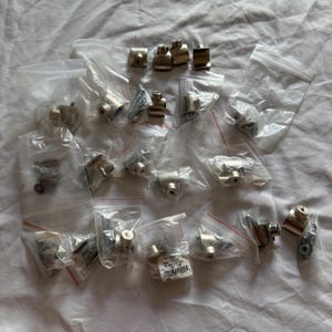 Puede incluir: Una colección de pequeñas piezas de ferretería metálicas de color plateado, que incluyen varias formas de conectores, tornillos y sujetadores, están empaquetadas en bolsas de plástico transparente con cierres de cremallera rojos. El hardware parece nuevo y sin usar.