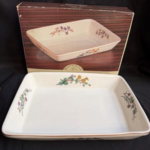 Puede incluir: Fuente rectangular para hornear de color crema con detalles florales y un borde marrón. La fuente se muestra frente a su caja original, que también presenta una imagen similar de la fuente. La caja tiene la palabra "WOODHILL" impresa.