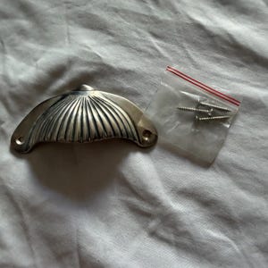 May include: A silver-toned, shell-shaped drawer pull with a ridged design. The pull has two holes for mounting and is accompanied by a small bag of screws. The hardware is likely for furniture or cabinetry.