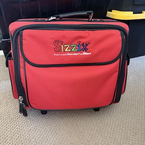 May include: A red rolling craft bag with black trim and a black handle. The bag features the Sizzix logo and a zippered front pocket. The bag is on a beige carpet.