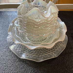 Op de afbeelding: Een stapel iriserende glazen kommen en borden. De kommen hebben een gerimpelde rand en een golvend ontwerp. De borden hebben een textuur met een bubbelachtig patroon. Het glas heeft een parelmoerachtige, opalescerende glans.