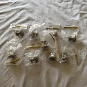 May include: A collection of cabinet knobs in clear plastic packaging. The knobs are a muted silver color with a decorative design. Each knob is paired with a gold-toned screw. The knobs are arranged on a white fabric surface.