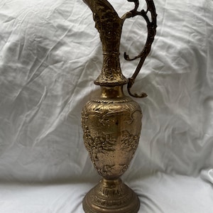 Könnte beinhalten: Eine goldfarbene Metallkanne mit einem verzierten Design und einem dekorativen Griff. Die Kanne hat einen breiten Boden und einen schmalen Hals.