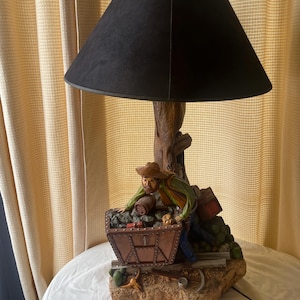 May include: A decorative table lamp featuring a black lampshade and a detailed base depicting a miner pushing a cart filled with coal. The base is made of a stone-like material, and the lamp has a rustic aesthetic.