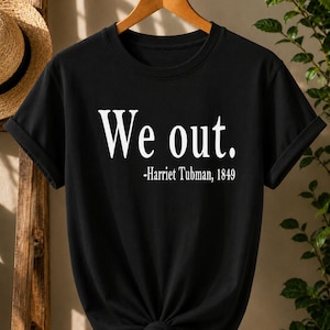 May include: A black short-sleeved t-shirt featuring white text that reads "We out." followed by "-Harriet Tubman, 1849" printed in the centre of the chest. The garment has a crew neckline and is displayed on a wooden hanger with rolled sleeves.