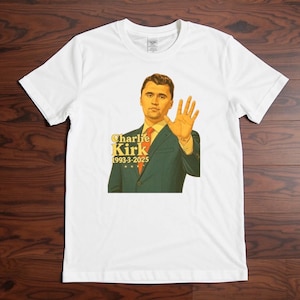 May include: White t-shirt featuring a graphic of a man in a suit with the text "Charlie Kirk 1993-3-2025". The man is holding up his hand. The graphic is in shades of yellow, red, and green.