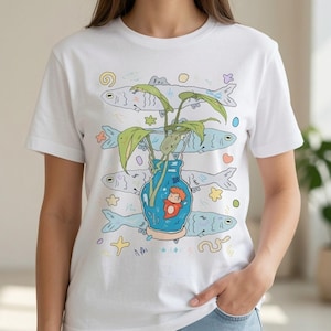 May include: A white short-sleeve t-shirt featuring a graphic print of a blue vase containing a green plant and a small orange creature. The design includes four light blue fish arranged horizontally behind the vase, surrounded by small colorful shapes.