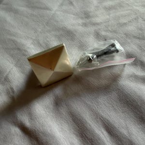 Puede incluir: Un tirador de armario cuadrado y geométrico con acabado metálico, que presenta un diseño triangular en tonos blancos y dorados. Incluye tornillos en una pequeña bolsa de plástico para la instalación. Herrajes para muebles.