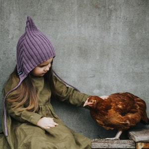 Op de afbeelding: Een jong meisje draagt ​​een paarse gebreide muts met lange banden en een groene linnen jurk. Ze zit op een houten bankje en aait een bruin kippetje.