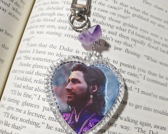 May include: A heart-shaped keychain with a clear plastic frame and a purple amethyst charm. The keychain features a printed image of a man with dark hair and a beard, wearing a purple and black outfit.