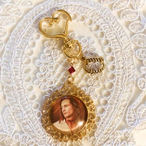 May include: A gold-toned keychain featuring a heart-shaped clasp, a small red crystal accent, and a charm shaped like a set of vampire fangs. A circular pendant with an ornate filigree frame displays a portrait of a person with long hair.