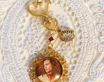 Puede incluir: Un llavero dorado con un cierre en forma de corazón, un pequeño cristal rojo y un dije con forma de colmillos de vampiro. Un colgante circular con un marco de filigrana ornamentado muestra el retrato de una persona de pelo largo.