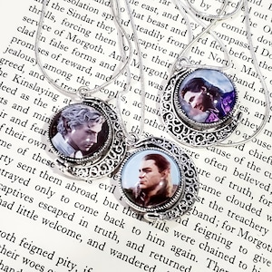 May include: Three silver necklaces with crescent moon pendants. Each pendant features a different image of a man with dark hair and a beard. The necklaces are on a book with text about Morgoth.
