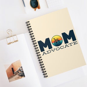 May include: A spiral-bound notebook with the words "MOM ADVOCATE" in navy blue. The "O" in "MOM" features a wheelchair graphic set against a sunset design. The notebook is cream-colored and rests on a white surface.