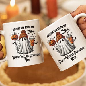 May include: Two white ceramic mugs with black handles. The mugs feature a cartoon ghost wearing a beanie hat and holding a pumpkin and a cup of coffee. The text on the mugs reads "Nothing can scare me" and "Dandy-Walker Syndrome" with "Dad" on one mug and "Mom" on the other.