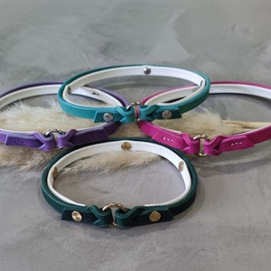 Op de afbeelding: Een collectie van vier leren halsbanden in verschillende kleuren: paars, turquoise, roze en groen. Elke halsband heeft een witte binnenvoering, een metalen sluiting en decoratieve stiksels. Halsbanden voor huisdieren.