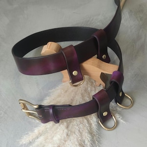 May include: Two leather belts with gold hardware. The larger belt is purple and black with a buckle. The smaller belt is purple with two D-rings.