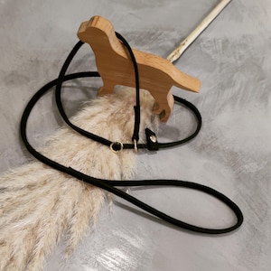 Op de afbeelding: Een zwarte leren hondenriem met gouden beslag. De riem is om een houten hondenfiguur gewikkeld.