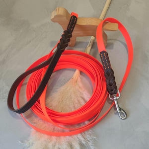 Op de afbeelding: Fel oranje hondenriem met een donkerbruin leren handvat en een zilverkleurige metalen sluiting. De riem is opgerold en ligt naast een houten honden-vormige decoratie.