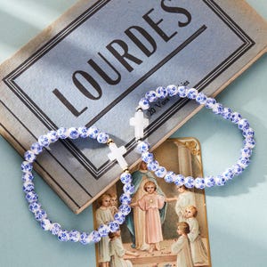 May include: Two white ceramic bracelets with blue floral designs and gold accents. Each bracelet features a white cross charm. The bracelets are displayed on a book with the word "Lourdes" printed on the cover.