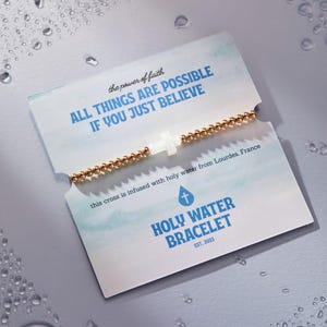 May include: A gold beaded bracelet with a white cross charm, displayed on a card with the text "ALL THINGS ARE POSSIBLE IF YOU JUST BELIEVE." The card also reads "HOLY WATER BRACELET EST. 2023."