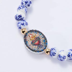 May include: A beaded bracelet featuring white and blue floral patterned beads and gold-colored accents. The focal point is an oval charm with a heart design, surrounded by a colorful stained-glass effect.