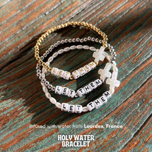 May include: Three layered bracelets with white beads and gold and silver beads. The bracelets spell out "Let Them" and feature a small white cross charm. The bracelets are infused with water from Lourdes, France. "Holy Water Bracelet" is written below the image.