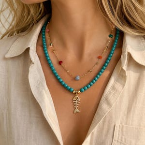 May include: A layered necklace set featuring a strand of turquoise-coloured beads with a gold-toned fish skeleton pendant encrusted with clear stones, and a delicate gold-toned chain accented with small, multicoloured beads and a central light blue bead.
