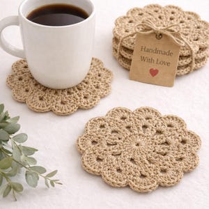 Può includere: Set di sottobicchieri all'uncinetto beige con un'etichetta con la scritta "Handmade With Love". Una tazza bianca con caffè è appoggiata su un sottobicchiere. I sottobicchieri hanno un diametro di circa 10 cm.