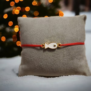 May include: Red string bracelet with a white fish charm featuring a blue evil eye. The bracelet is on a gray velvet cushion.