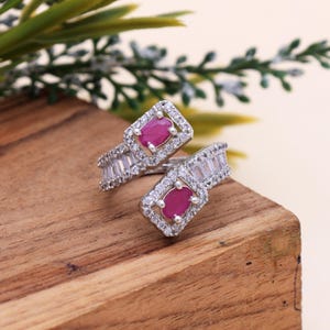 Op de afbeelding: Een zilverkleurige ring met twee ovale, magenta-kleurige edelstenen, elk omgeven door een vierkant van kleine, heldere stenen. De ringband heeft een rij rechthoekige, heldere stenen. De ring wordt gepresenteerd op een houten oppervlak.