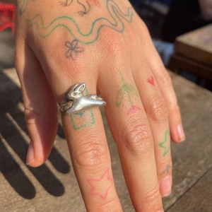 May include: A silver ring with a rabbit design. The ring is worn on the middle finger of a hand with several small tattoos.