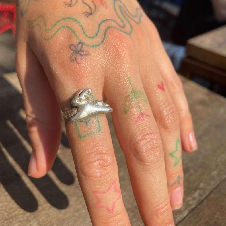 May include: A silver ring with a rabbit design. The ring is worn on the middle finger of a hand with several small tattoos.