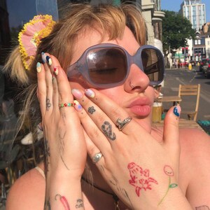 May include: A person with short blonde hair wearing blue tinted sunglasses and a nose ring. They have multiple tattoos on their hands, including a scorpion, a flower, and a red drawing of a creature. Their fingernails are painted with colorful designs.
