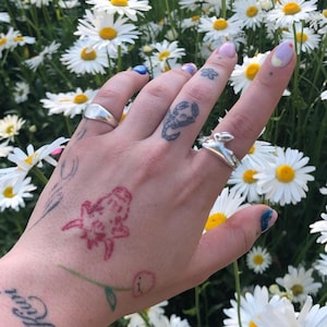 May include: A hand with several tattoos, including a scorpion, a flower, and a red creature, is wearing two silver rings. The hand is surrounded by white daisies.