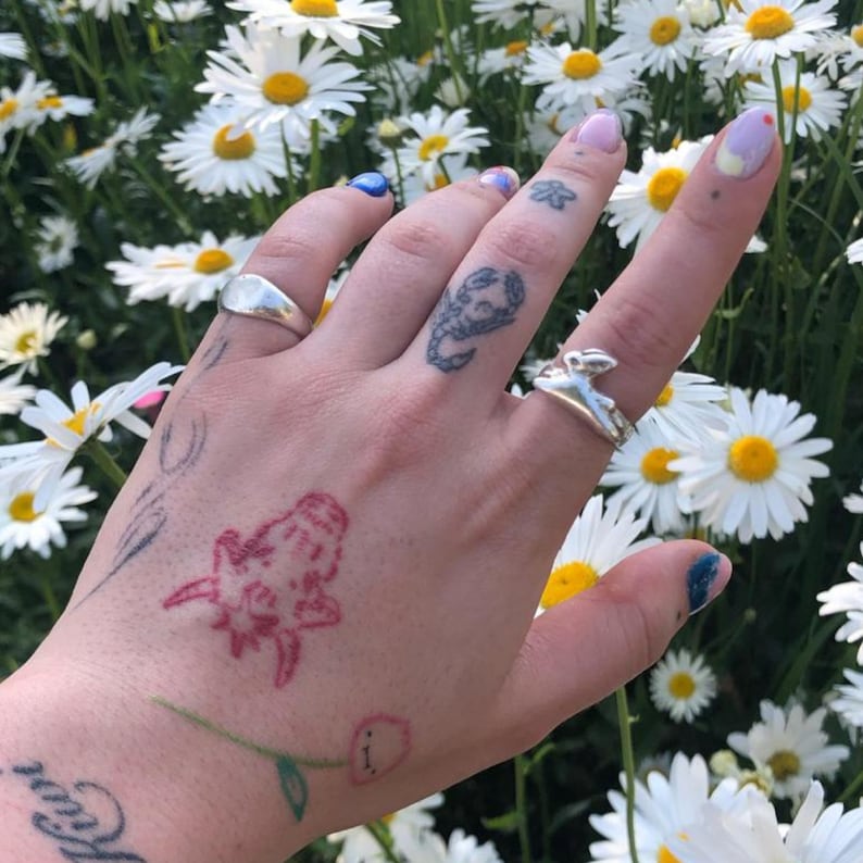 May include: A hand with several tattoos, including a scorpion, a flower, and a red creature, is wearing two silver rings. The hand is surrounded by white daisies.