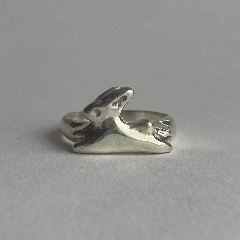 May include: A silver ring featuring a stylized rabbit design. The rabbit is depicted in a leaping pose, with its ears and tail visible.
